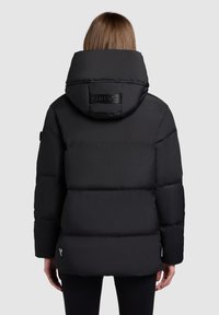 Zwarte pufferjack met een grote capuchon, geborduurd ontwerp en glanzende afwerking. Heeft een label op de rug en een slanke pasvorm.