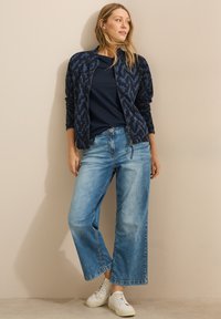Cazadora bomber navy con patrón geométrico texturizado, combinada con jeans de pierna ancha en azul claro y zapatillas blancas.