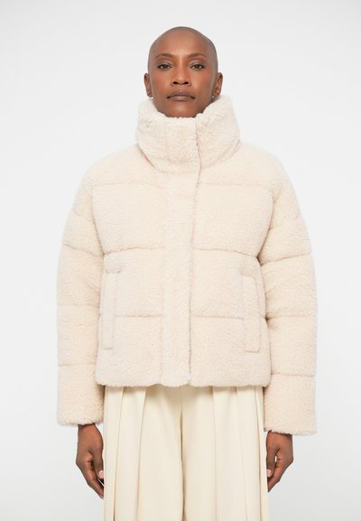 Veste en quilted de couleur crème, moelleuse, avec un col haut, des poches avant et une silhouette carrée. Présente une texture douce et un design minimaliste.