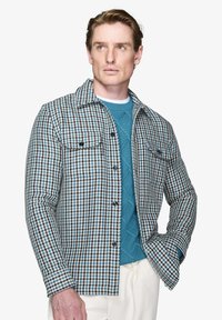 Mønstret skjortejakke i dæmpet blå, hvid og sort houndstooth; har to brystlommer og knaplukninger. Bæres over en tekstureret sweater.