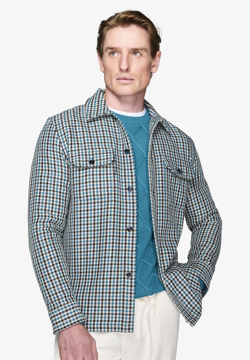 Mønstret skjortejakke i dæmpet blå, hvid og sort houndstooth; har to brystlommer og knaplukninger. Bæres over en tekstureret sweater.