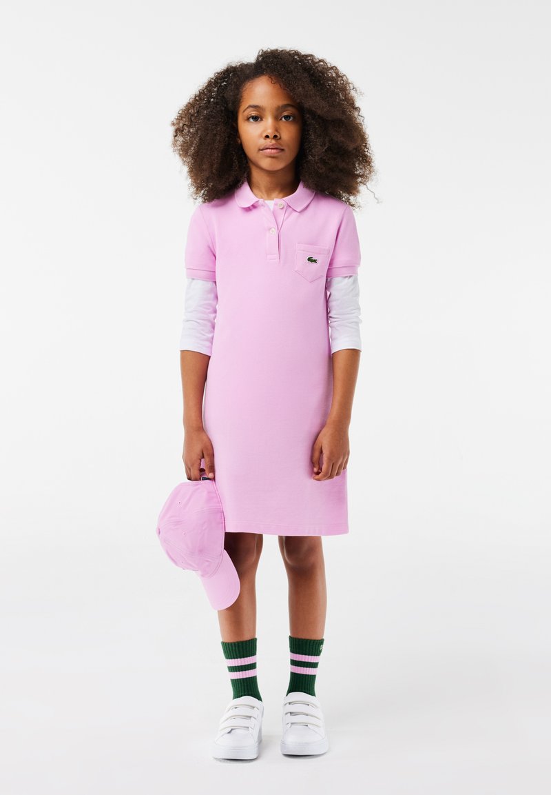 Vestido polo rosa de manga corta, que cuenta con un bolsillo y un logo bordado. Combinado con una gorra a juego, calcetas a rayas verdes y zapatillas blancas.