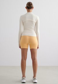 Lys beige langermet topp med en tettsittende design, kombinert med oransje sykkelshorts. Glatt stoff og minimale sømmer, brukt med hvite joggesko.