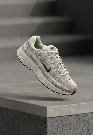 Beige en bruine Nike hardloopschoen met slangenvelpatroon, zwarte swoosh en dikke zool, hangend boven een betonnen oppervlak met trappen op de achtergrond.