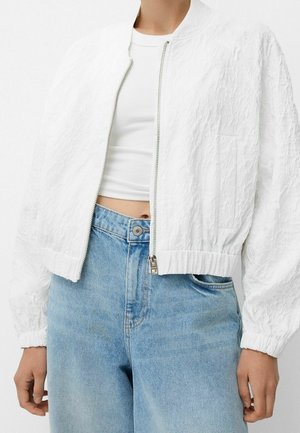 Witte, gestructureerde ritsjas die open gedragen wordt over een wit geribd crop topje, gecombineerd met lichtblauwe, hoogwater spijkerbroek met hoge taille.