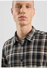 Camicia di flanella con bottoni, in un motivo a quadri nero, marrone e bianco. Presenta un colletto e due bottoni visibili all'altezza del colletto.