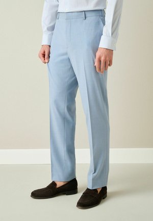 Man wearing light blue dress trousers, white shirt, and dark brown suede loafers standing against neutral background.