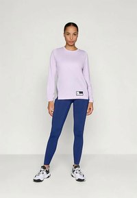 Long-sleeve light purple top with a round neckline, paired with blue leggings. Black and white logo patch on the top's lower left side.