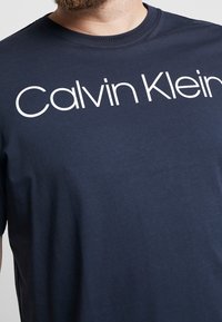 Mies, joka käyttää tummansinistä T-paitaa, jossa on suuri valkoinen "Calvin Klein" -logo rintaan, leikattu nenän alapuolelta.