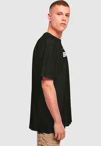 Black cotton t-shirt with short sleeves, crew neckline, and front graphic text. Loose fit, reaching mid-hip, with a smooth texture.