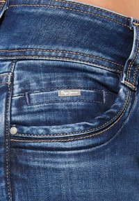 Close-up stražnjeg džepa na plavim trapericama s malom oznakom "Pepe Jeans" i kontrastnim narančastim šavovima.