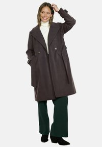 Cappotto viola scuro con ampi rever, bottoni doppi e tasche laterali. Abbinato a un dolcevita bianco e pantaloni verdi.
