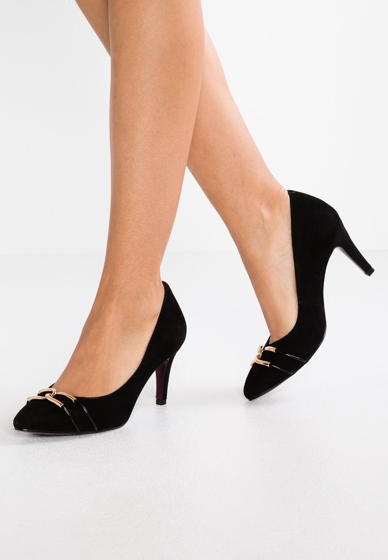 Sorte ruskind højhælede sko med spids tå, der har en guldtonet hardwaredetalje og en blank sort detalje over fronten.
