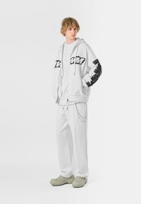 Sudadera blanca con cremallera y detalles en negro, combinada con pantalones blancos a juego que presentan un detalle de cadena. Llevado con calzado texturizado de color verde claro.