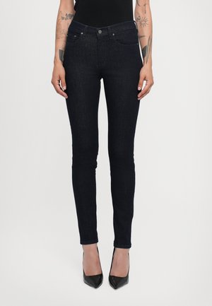 Kobieta ubrana w czarne obcisłe dżinsy i czarne wysokie obcasy, stojąca z ramionami swobodnie opuszczonymi wzdłuż ciała na białym tle.