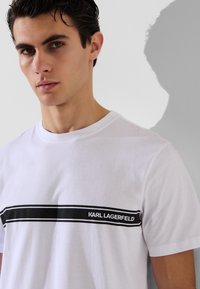 T-shirt bianco in cotone con una striscia orizzontale nera e la scritta "KARL LAGERFELD", scollo a girocollo, maniche corte, texture liscia.
