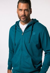 Teal zip-up hoodie med dragsko, framfickor och broderad logotyp. Tillverkad av mjukt tyg, med en avslappnad passform och långa ärmar.