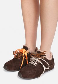 Brune semskede slip-ons med pelsfôr, brukt uten sokker, med uliksnoring: en oransje og en hvit med svarte striper.