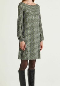 Robe verte à motifs avec manches longues, col rond et coupe ample. Présente un design géométrique avec des accents violets. Portée avec des bottes noires montant jusqu'aux genoux.