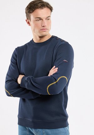 Homme aux bras croisés, portant un pull bleu marine avec des boutons aux épaules et des coudières jaunes, regardant sur le côté.