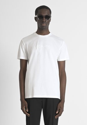 Camiseta blanca de algodón con cuello redondo, mangas cortas y un logo en relieve en el pecho; combinada con pantalones oscuros y gafas de sol.