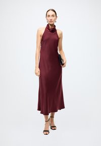 Robe en satin bordeaux avec un décolleté bénitier et une silhouette fluide. Le mannequin tient une pochette noire et porte des talons noirs à brides.