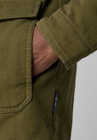 Main placée dans la poche d'une veste texturée vert olive avec une étiquette visible "EST. 1993" sur la couture latérale.
