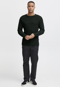 Knitted dark green sweater with a round neckline, textured fabric, and long sleeves. Paired with black pants and dark sneakers.
