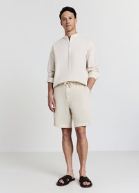 Light beige, long-sleeve henley shirt with button placket; matching, knee-length shorts with drawstring; black slip-on sandals.