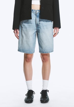 Person iført lyseblå denimshorts, hvide sokker, sorte snørestøvler, en sort jakkesætjakke med nålestriber og ringe på begge hænder.