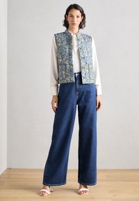 Γαλάζιο γιλέκο με λουλουδάτο μοτίβο, υψηλό λαιμό και κουμπί, συνδυασμένο με παντελόνι από σκοτεινή τζιν και κρεμ σανδάλια.