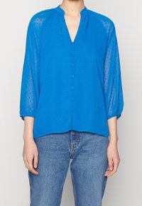 Blå skjorta med krage och V-hals, texturerat prickigt mönster, genomskinligt tyg och trekvartsärmar som är rynkade vid ärmsluten.