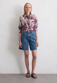 Blumenhemd mit Knopfverschluss in Lila und Creme, kombiniert mit blauen Denim-Shorts und braunen Sandalen. Glattes Material und lässige Passform.