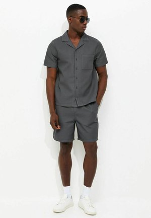 Mężczyzna ubrany w ciemnoszarą koszulę z krótkim rękawem zapinaną na guziki, dopasowane krótkie spodenki, białe trampki, skarpetki i czarne okulary przeciwsłoneczne stojący przy gładkiej białej ścianie.