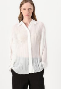 Blusa bianca plissettata con colletto a camicia, caratterizzata da un effetto sfumato che va dal bianco al grigio, e maniche lunghe con polsini arrotolati.