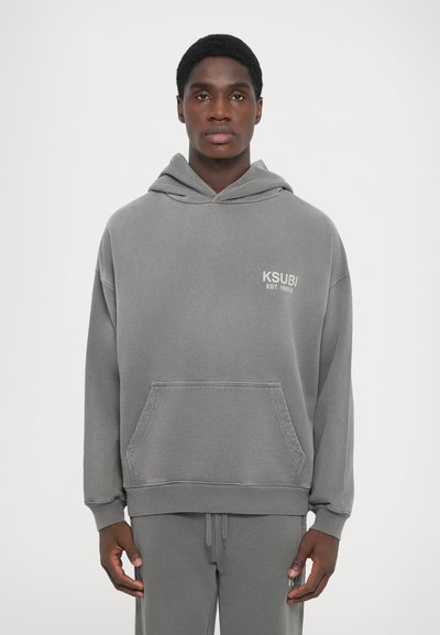 Gray oversized hoodie with a kangaroo pocket and drawstring hood. Features a white KSUBI logo on the chest. Soft cotton material.