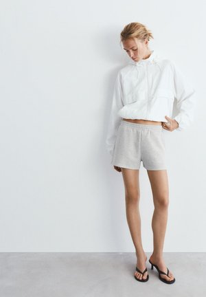 Frau trägt weiße kurze Jacke, graue Shorts mit elastischem Bund und schwarze Sandalen mit Absatz, steht vor einer weißen Wand.