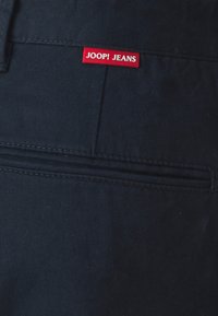 Tumši zili audumi ar gludu tekstūru, ar aizmugurējo kabatu un sarkanu "JOOP! JEANS" birku, kas iešūta jostasvietā.