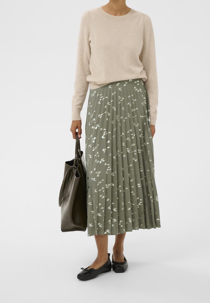 Femme portant un pull beige et une jupe plissée verte avec des motifs blancs, des ballerines noires, tenant un grand sac cabas vert foncé.