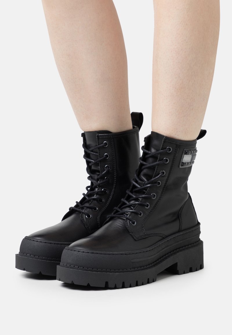 Bottes de combat noires à lacets avec semelles épaisses en caoutchouc portées sur des jambes nues sur un fond clair uni.