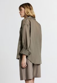 Donna con capelli biondi di lunghezza spalla che indossa una camicia oversize verde oliva e pantaloni shorts larghi color taupe, in piedi di profilo contro uno sfondo neutro.
