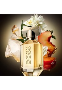 Botella de fragancia con un diseño de vidrio facetado, acentos dorados y una tapa cuadrada. Rodeada de flores melocotón, flores blancas y elementos que parecen cristal.