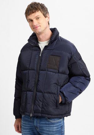 Marineblauer Pufferjacke mit gestepptem Design, Frontreißverschluss, hohem Kragen und schwarzem Logoauflage. Verfügt über elastische Bündchen und seitliche Taschen.