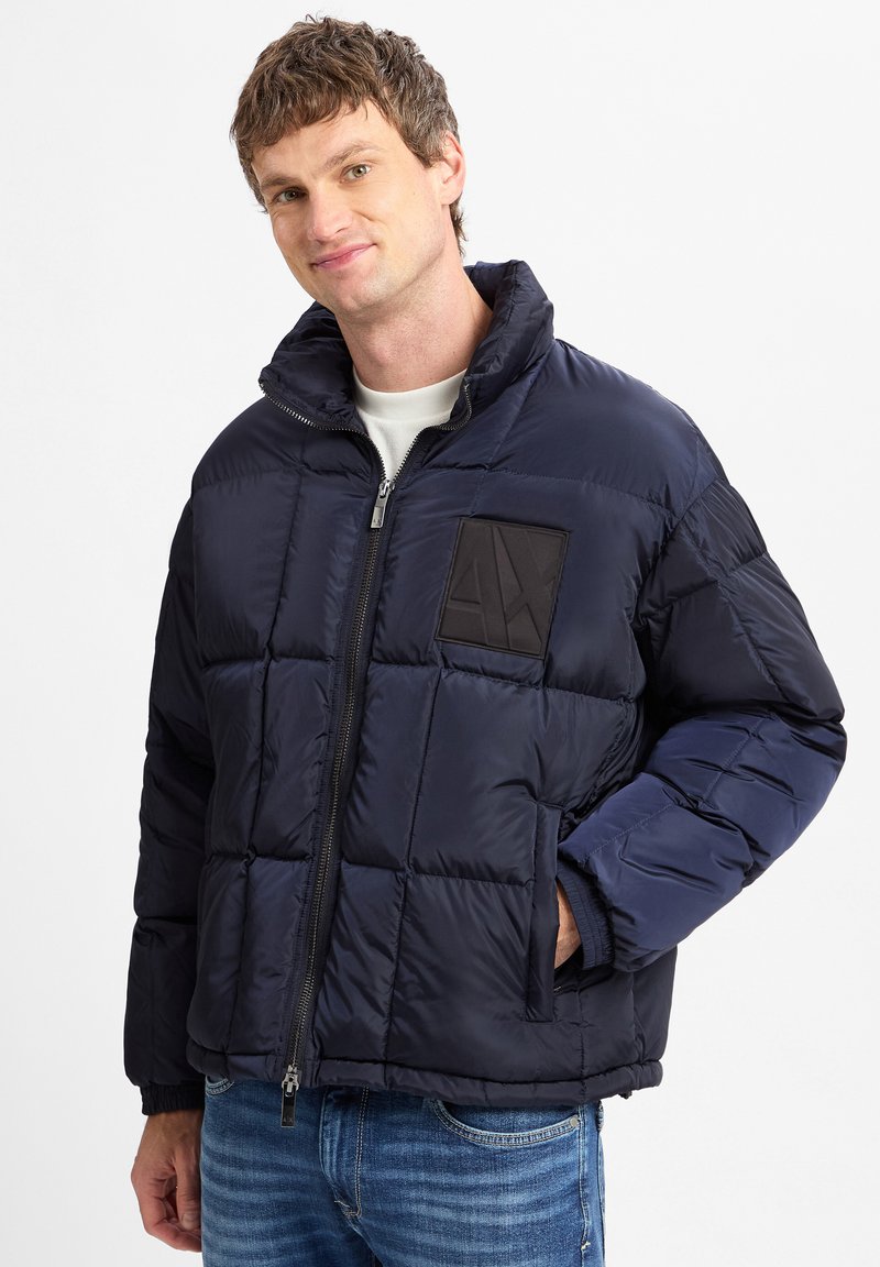Giubbino imbottito blu navy con design trapuntato, zip frontale, colletto alto e un'etichetta logo nera. Presenta polsini elasticizzati e tasche laterali.