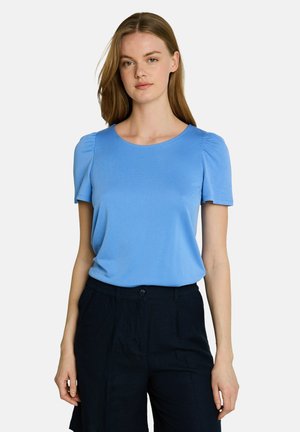 Junge Frau mit glatten hellbraunen Haaren, die ein hellblaues Kurzarmshirt und schwarze Hose vor weißem Hintergrund trägt.