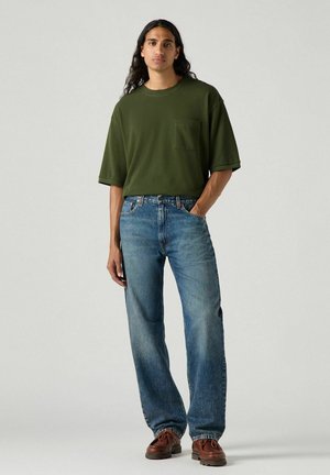 Groene t-shirt met korte mouwen en een borstzak, gecombineerd met lichtblauwe spijkerbroek. Het model staat in bruine schoenen op een grijze achtergrond.