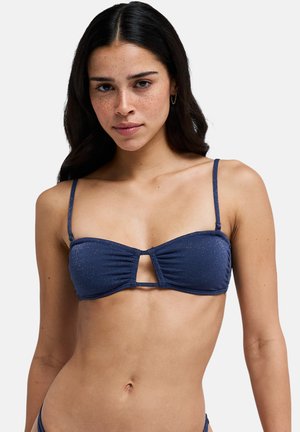 Młoda kobieta o długich ciemnych włosach i piegach, ubrana w granatowy top bikini z cienkimi ramiączkami i małym wycięciem z przodu.