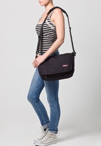 Borsa a tracolla nera realizzata in tessuto resistente, con un flap pieghevole, una tracolla regolabile e una patch con logo. Texture liscia con dettagli minimi.