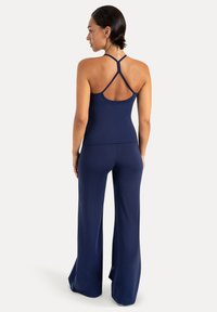 Femme vue de dos portant un top bleu marine sans manches avec des bretelles croisées et un pantalon large assorti sur un fond uni.