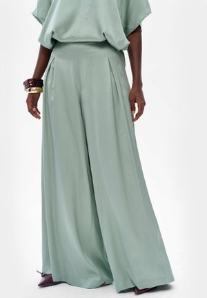 Pantalons larges et fluides en tissu vert menthe doux, avec des plis, une texture lisse et une taille haute. Complétés par des bracelets empilés.